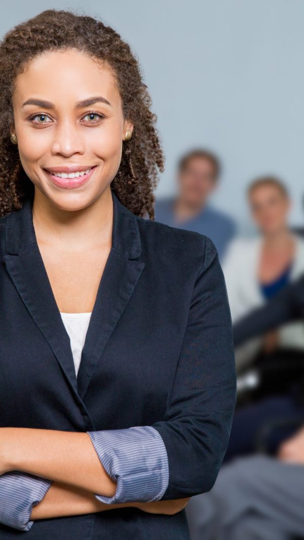 Portrait of female African American executive manager standing in meeting room and smiling, business people sitting at office desk on background